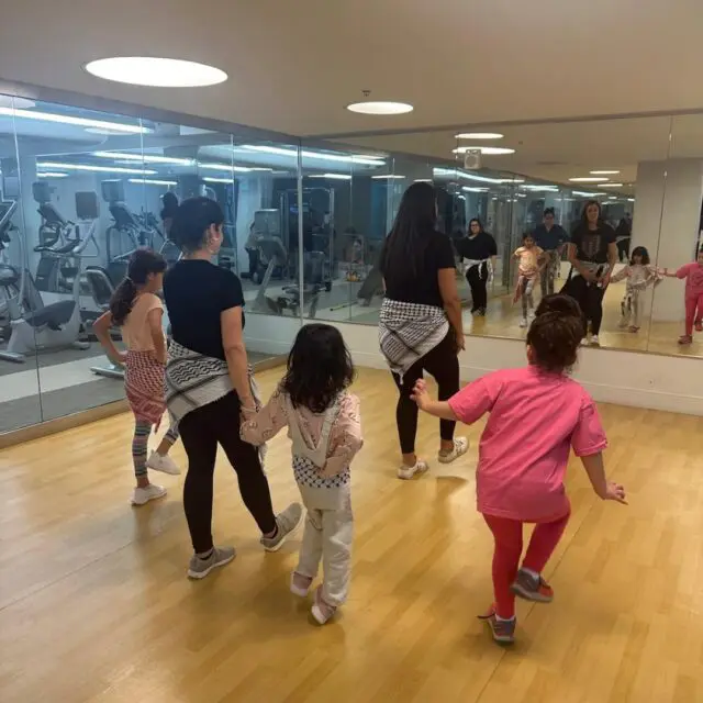 Group of children singing into microphones during a music class while an instructor plays an instrument, with a grand piano in the background.