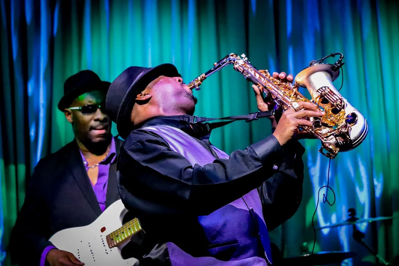 Saxophonist passionately performing on stage with a guitarist in the background under colorful stage lighting.